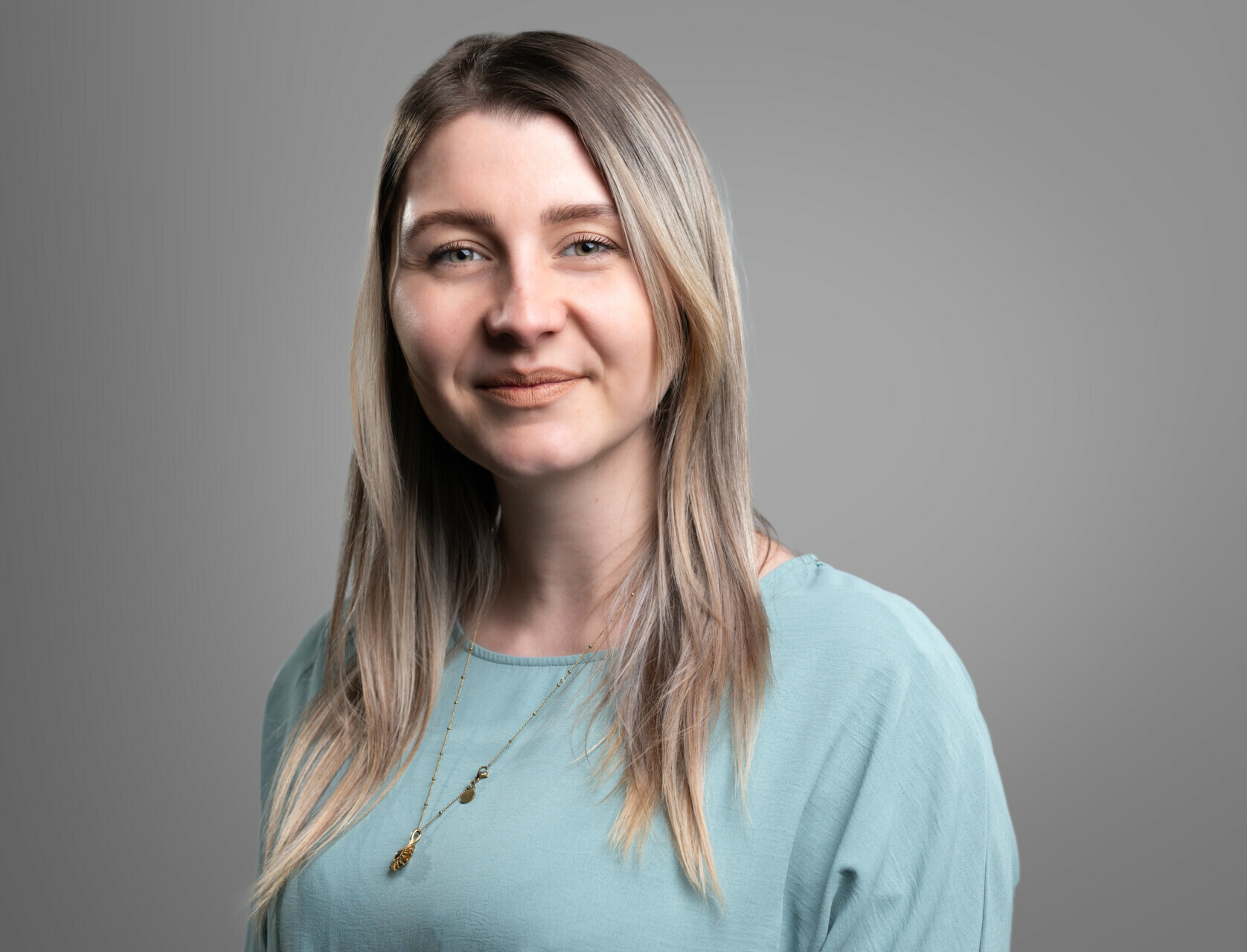 Eine lächelnde Frau mittleren Alters mit langem, glattem, hellbraunem Haar, einem türkisfarbenen Oberteil und einer schlichten Halskette steht vor einem grauen Hintergrund.