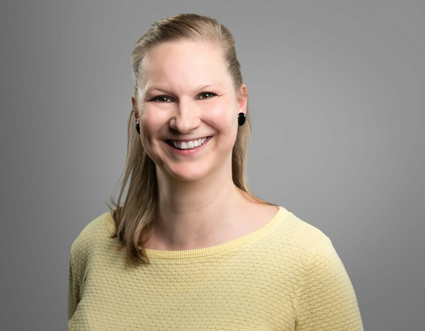 Ein Porträt einer lächelnden Frau mit blonden Haaren, die ein gelbes Oberteil trägt und vor einem grauen Hintergrund steht.
