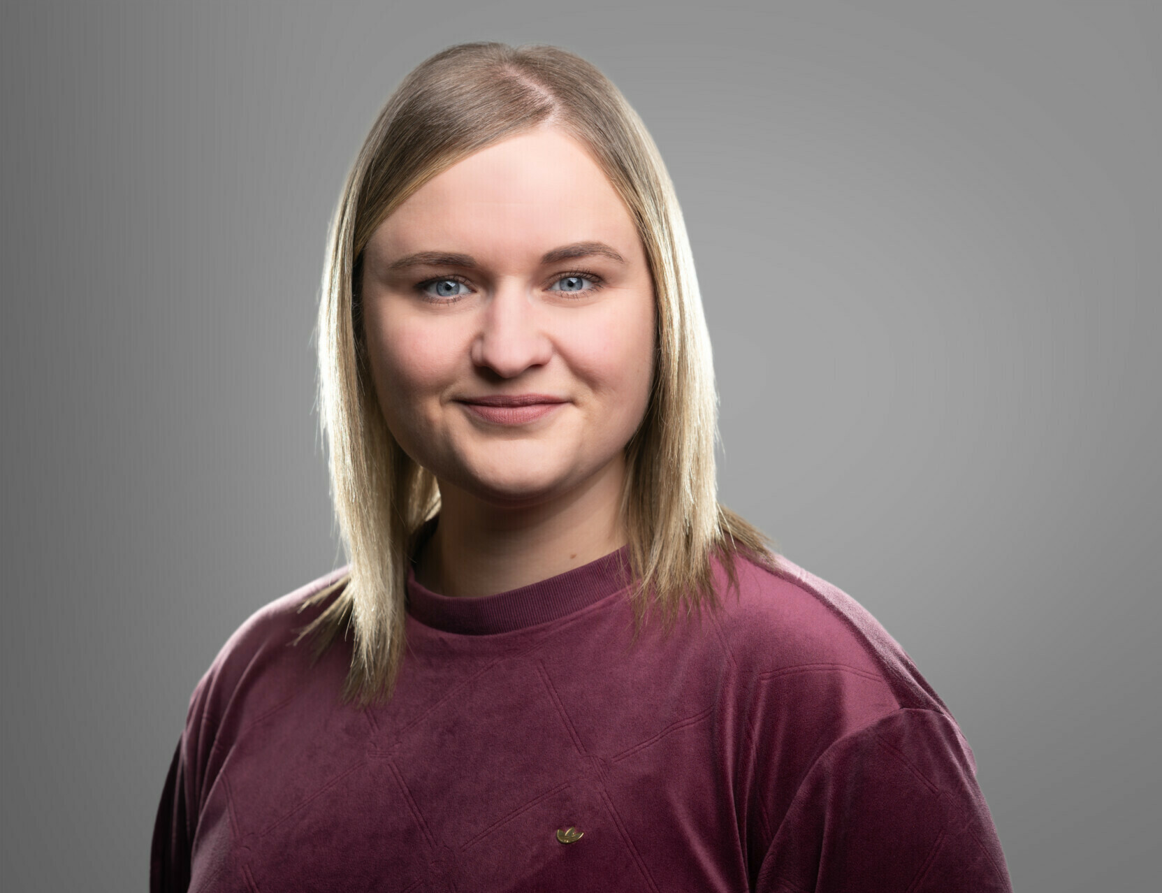 Ein Porträt einer lächelnden Frau mit mittellangem blondem Haar und einem dunkelroten Oberteil vor einem grauen Hintergrund.