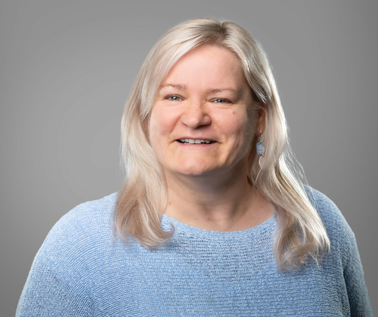 Porträt einer lächelnden Frau mittleren Alters mit blonden Haaren, die einen hellblauen Pullover trägt, vor einem grauen Hintergrund.