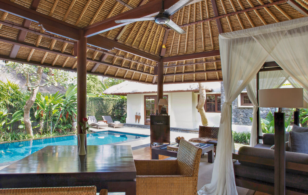 Ein luxuriöses, offen gestaltetes Wohnzimmer mit strohgedeckter Decke, das nahtlos in einen Außenbereich mit privatem Swimmingpool übergeht. Rattanmöbel und ein weißer Vorhang, der einen gemütlichen Sitzbereich schafft, sorgen für ein tropisches Ambiente. Im Hintergrund ist üppige Vegetation zu sehen.