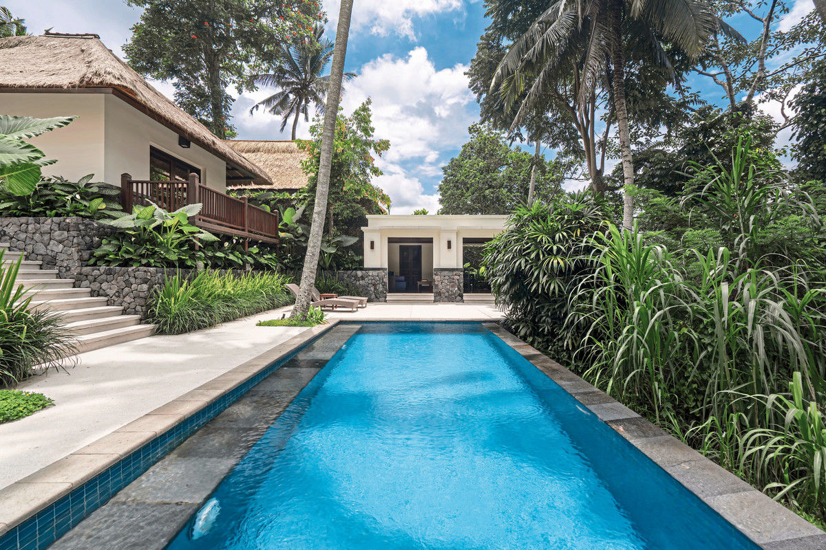Ein luxuriöser Swimmingpool mit klarem, blauem Wasser, umgeben von üppiger tropischer Vegetation und Palmen. Rechts im Bild befindet sich ein gepflegter Garten. Am Ende des Pools steht ein stilvolles, weißes Gebäude mit offenstehenden Türen, das eine einladende Atmosphäre erzeugt. Auf der linken Seite des Bildes sieht man ein traditionelles Haus mit Strohdach und einer Holzterrasse.
