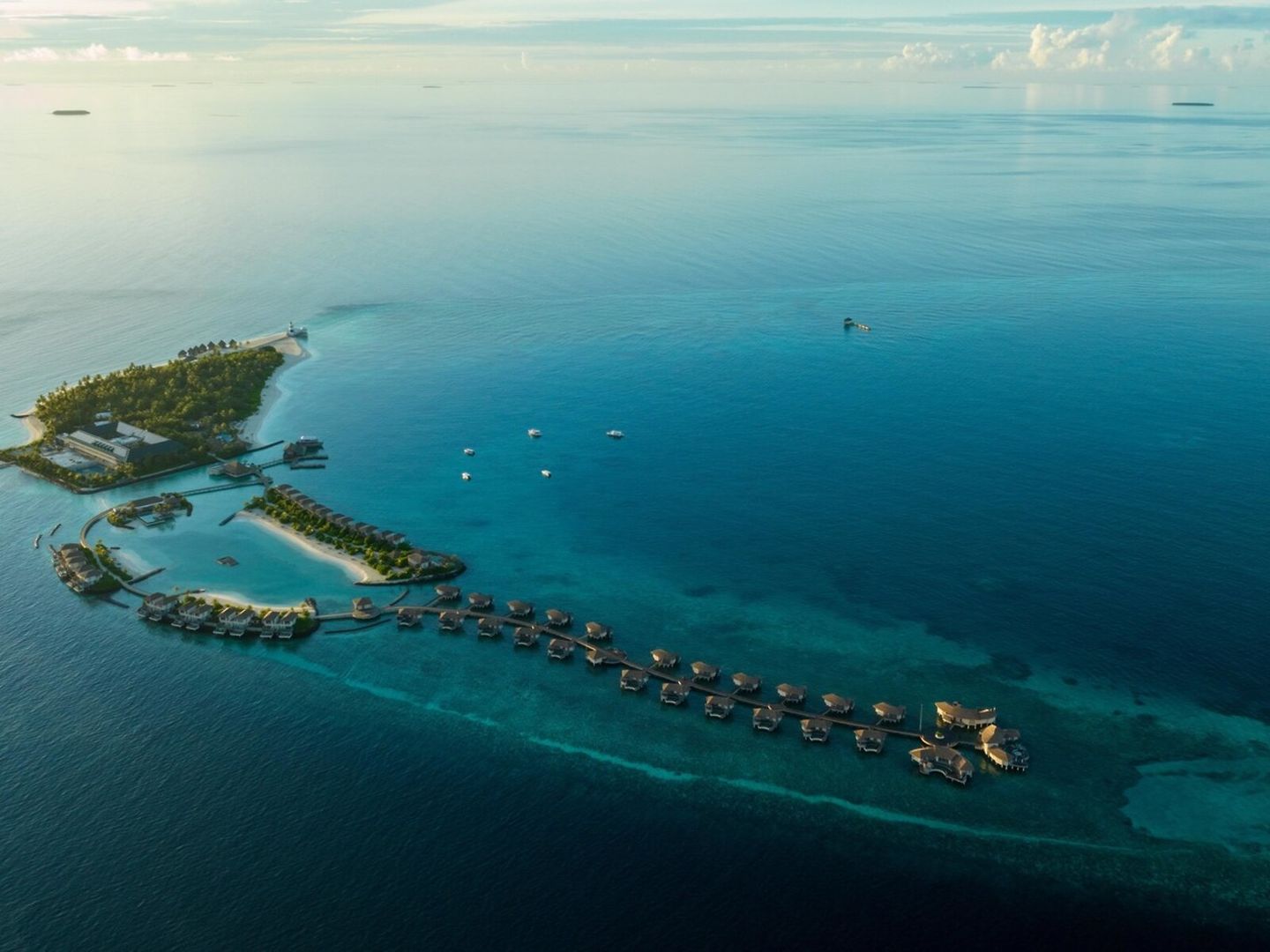 Resortüberblick Luftaufnahme einer tropischen Inselresort mit mehreren Überwasser-Bungalows und einem Leuchtturm, umgeben von ruhigem, klarem blauem Meerwasser und sichtbaren Korallenriffen bei Sonnenuntergang.