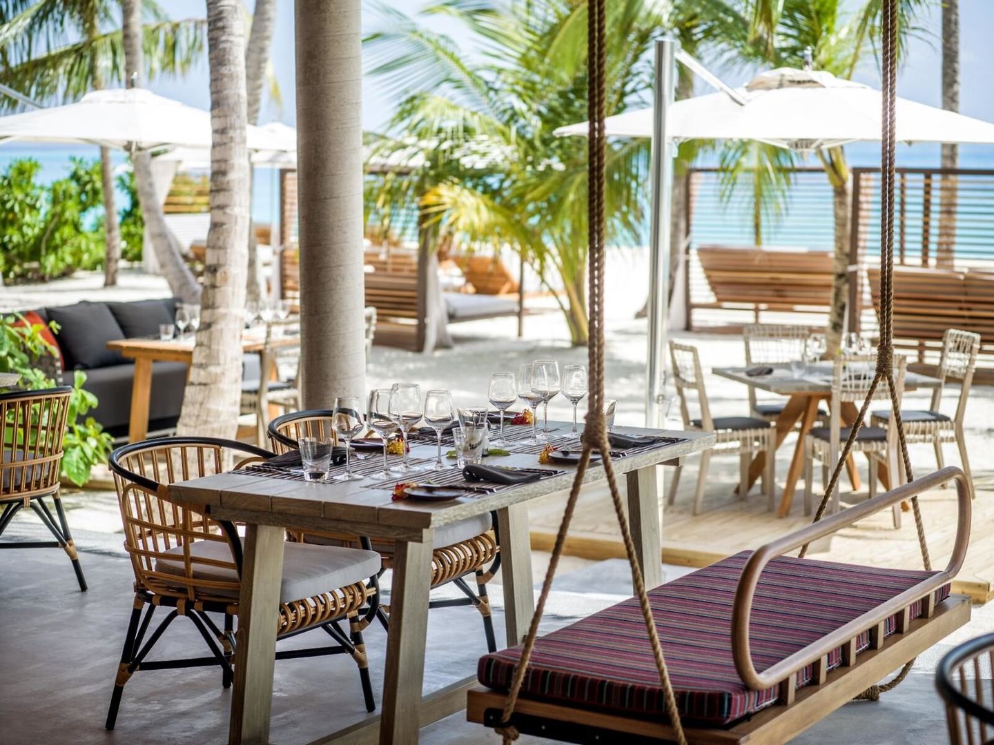 Restaurant Außenbereich eines eleganten Strandrestaurants mit hölzernen Esstischen und bequemen Sitzgelegenheiten unter Palmen. Ein Gedeck für mehrere Personen ist vorbereitet mit Gläsern und Besteck, und im Hintergrund sind Sonnenschirme und weitere Sitzbereiche zu sehen. Die entspannende Atmosphäre wird durch sanftes Tageslicht und das nahe Meer verstärkt.