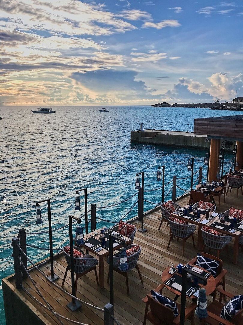 Restaurantterrasse Ein idyllisches Restaurant mit Holzterrasse direkt am Wasser unter einem beeindruckenden Himmel während des Sonnenuntergangs. Mehrere Tische sind elegant gedeckt und mit Laternen dekoriert. Im Hintergrund sind das ruhige Meer und einige Boote zu sehen.