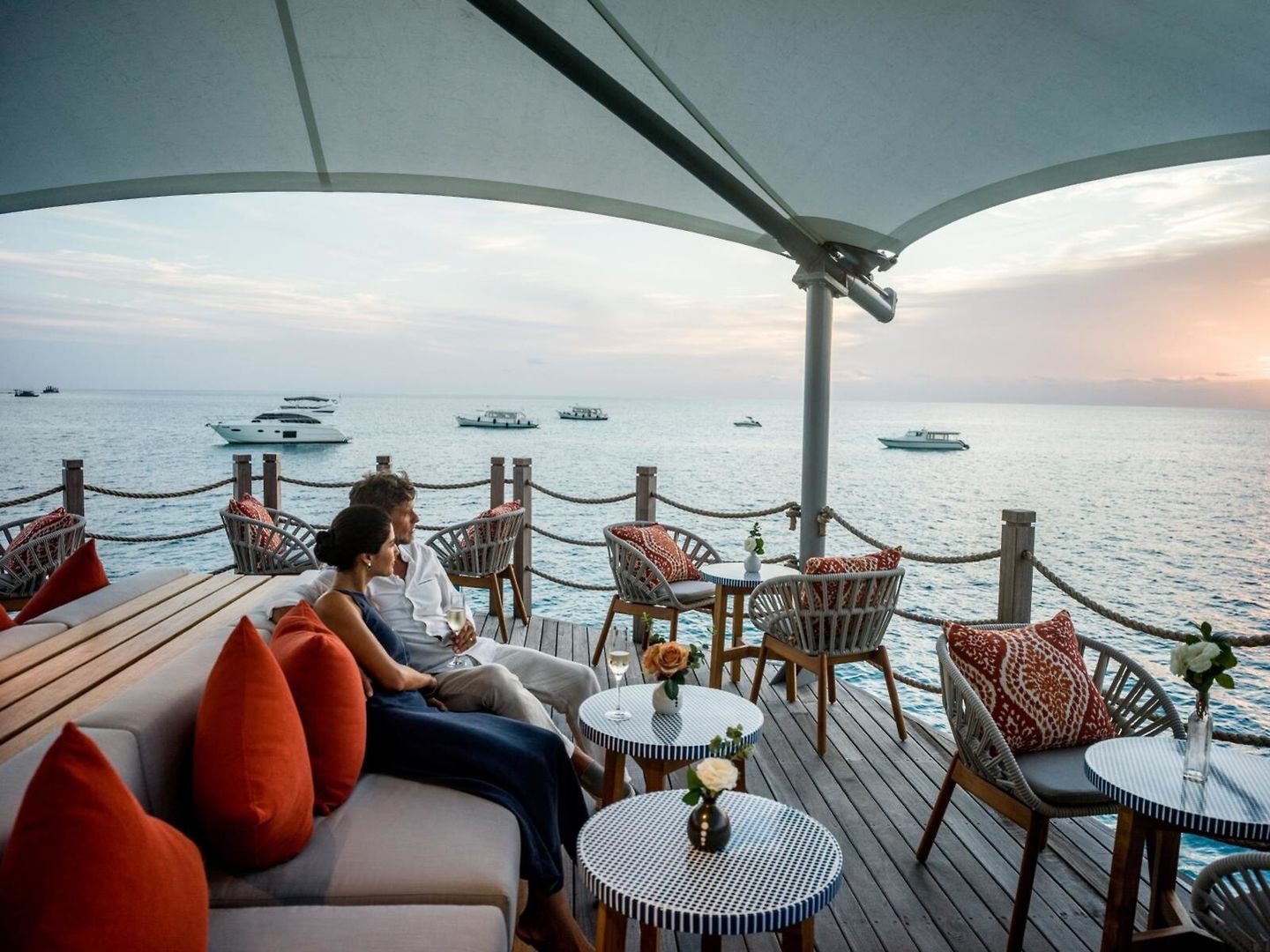 Romantik Ein Paar entspannt auf einer elegant eingerichteten Terrasse am Meer bei Sonnenuntergang. Sie sitzen auf einem Sofa unter einem schattenspendenden Sonnenschirm, umgeben von stilvollen Möbeln und dekorativen Kissen, mit Blick auf den ruhigen Ozean, gegenüber mehreren Yachten, die sanft auf dem Wasser schaukeln.