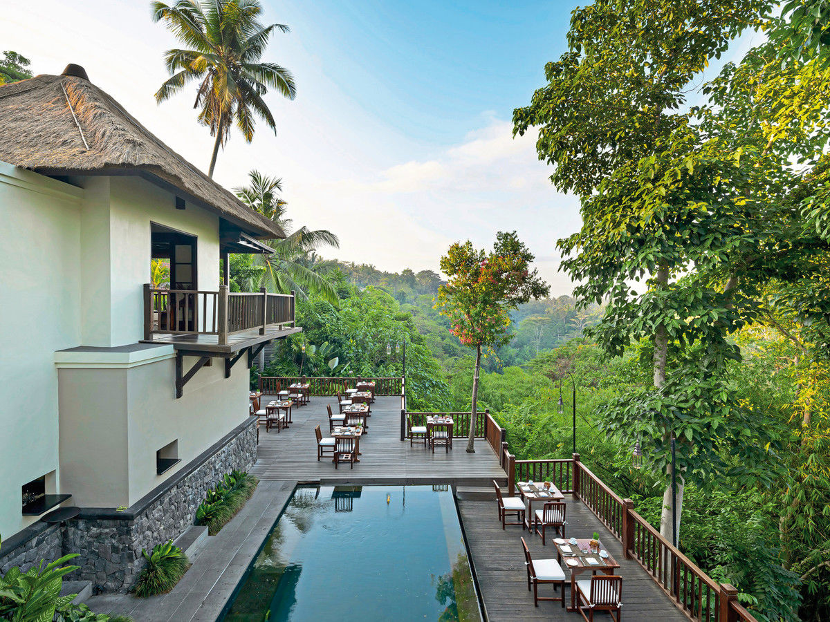 Terrasse im Ubud Private Villa & Spa-Pool Ein luxuriöses Resort mit einem offenen Speisebereich am Rande eines ruhigen Infinity-Pools überblickt ein üppiges, tropisches Waldtal. Traditionelle Architekturmerkmale werden bei der Gestaltung der weißen Gebäude mit Strohdächern deutlich. Elegante Tische sind für ein Essen im Freien vorbereitet.