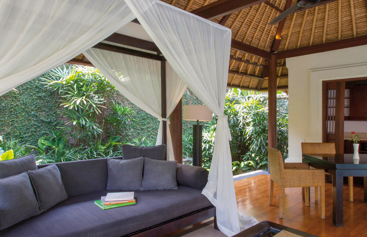 Alt-Text: Eine luxuriöse Außenlounge mit einem bequemen Sofa, umgeben von weißen Vorhängen, mit Blick auf einen üppigen grünen Garten. Das Interieur eines offenen Wohnraumes mit einem Esstisch aus Holz ist im Hintergrund zu sehen.