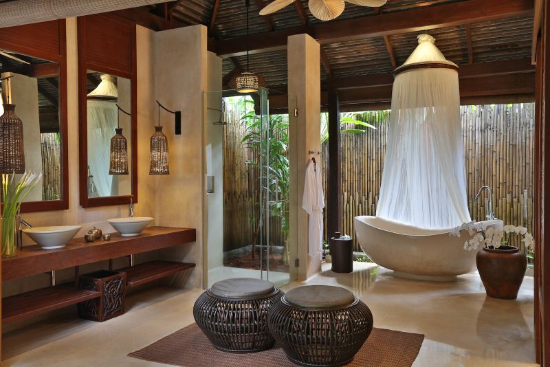 Badezimmer der Ocean Pool Suite Ein luxuriöses Badezimmer mit natürlichem Dekor, Holzakzenten und einer freistehenden Badewanne unter einem Moskitonetz. Zwei Waschbecken sitzen auf einer hölzernen Eitelkeit gegenüber von Spiegeln, akzentuiert durch hängende Pendelleuchten. Im Hintergrund führt eine Tür zu einer Duschkabine, abgeschirmt durch Bambuswände.