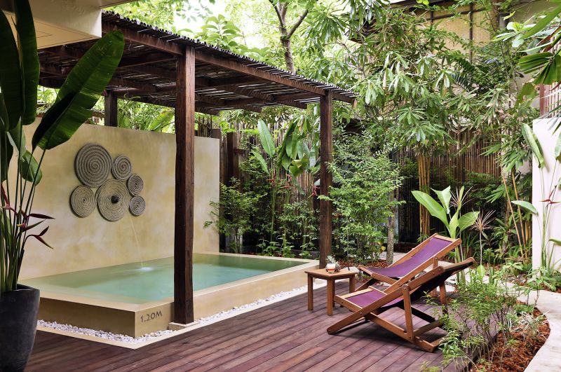 Garden Pool Suite Ein friedvoller Garten mit einem kleinen Pool, umgeben von üppigem Grün und einem Holzdeck. Auf der rechten Seite befindet sich eine braune Liegestuhl. An der Poolwand hängt ein dekoratives Kunstwerk aus Kreisen.