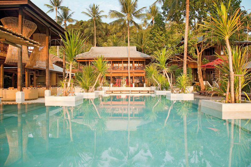 Infinitypool Ein luxuriöses Resort mit einem ruhigen Swimmingpool im Vordergrund, umgeben von Palmen und tropischen Pflanzen. Im Hintergrund befindet sich ein elegantes Gebäude im traditionellen Stil mit einer Holzfassade und weitläufigen Terrassen. Die Szene wird durch das warme Licht eines frühmorgendlichen oder spätnachmittäglichen Sonnenscheins akzentuiert, das eine friedliche und einladende Atmosphäre schafft.