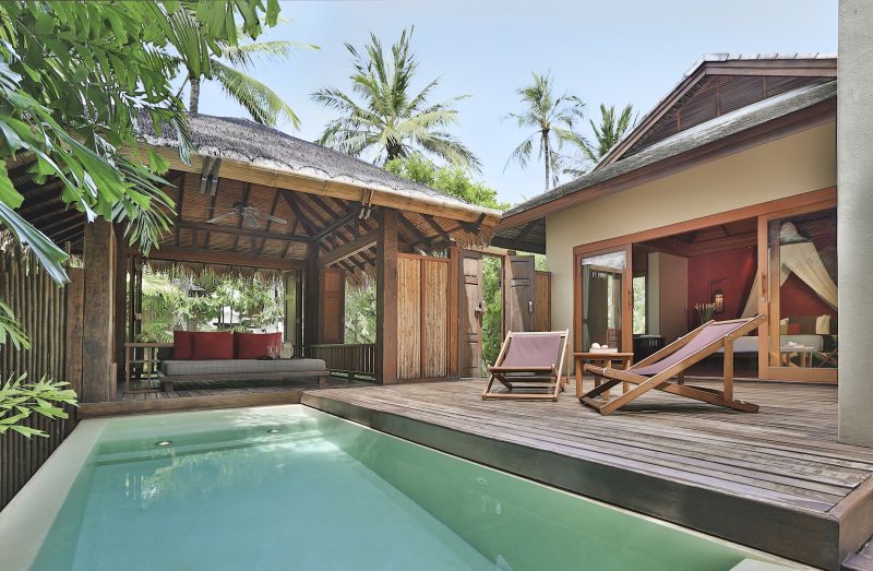 Lagoon Pool Suite Ein exklusives tropisches Resort mit einem einladenden Pool im Vordergrund, flankiert von Liegestühlen und umgeben von üppigem Grün und Palmen. Am Poolrand befindet sich ein offenes hölzernes Pavillon mit komfortabler Lounge-Ausstattung und einem Blick in den angelegten Garten. Im Hintergrund ist ein Teil des eleganten Ferienhauses mit offenstehenden Türen, durch die das Innere des Schlafzimmers mit einem großen Bett zu sehen ist.