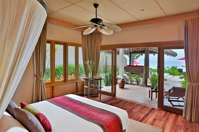 Ocean Garden Pool Suite Ein luxuriöses Schlafzimmer eines Resorts mit Blick auf das Meer durch große Glasschiebetüren. Ein großes Bett mit weißer Bettwäsche und einer roten Überwurfdecke steht im Raum, flankiert von eleganten Vorhängen und einem Deckenventilator. Die Einrichtung ist in erdigen Tönen gehalten, und draußen auf der Terrasse sind komfortable Liegestühle zu sehen, die eine entspannende Atmosphäre schaffen.