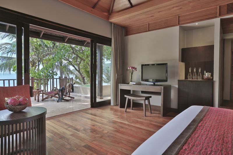 Ocean Pool Suite Innenansicht eines stilvollen Hotelzimmers mit Holzboden und Decke, einem großen Doppelbett im Vordergrund und einem Flachbildfernseher auf einem Schreibtisch. Durch die bodentiefen Fenster sieht man eine Terrasse mit einem hölzernen Liegestuhl und üppigem Grün, das die Sicht auf das Meer verdeckt.