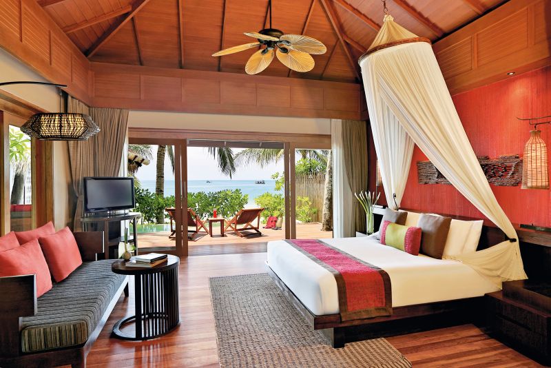 Ocean Pool Villa Ein luxuriöses Schlafzimmer in einem Strandresort mit offenen Schiebetüren zum Meer. Das Zimmer verfügt über ein großes Bett mit Moskitonetz, Holzböden und -decken, eine bequeme Sitzbank, einen Fernseher und ist von tropischer Natur umgeben.