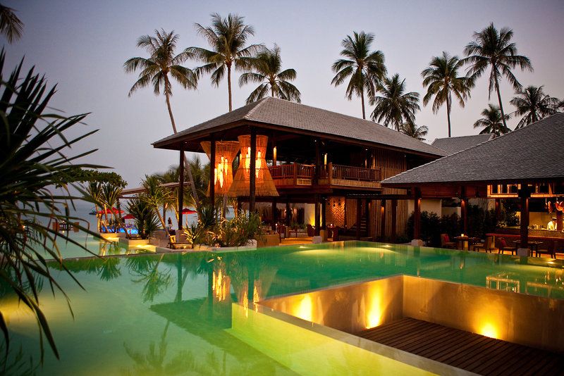 Pool am Abend Ein Luxus-Resort bei Dämmerung mit beleuchtetem Infinity-Pool im Vordergrund und traditionellen Holzbungalows, umgeben von Kokospalmen.