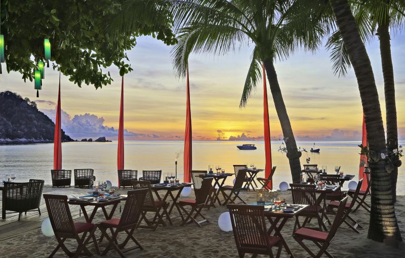 Restaurant The Bistro Ein elegantes Abendessen am Strand bei Sonnenuntergang mit arrangierten Tischen und Stühlen auf Sand. Rote Servietten und Tischdekorationen akzentuieren die Sitzgelegenheiten. Im Hintergrund sind Palmen, ein ruhiges Meer und der Himmel in warmen Farbnuancen von Gelb, Orange und Blau zu sehen. Einige Boote sind auf dem Wasser zu erkennen.