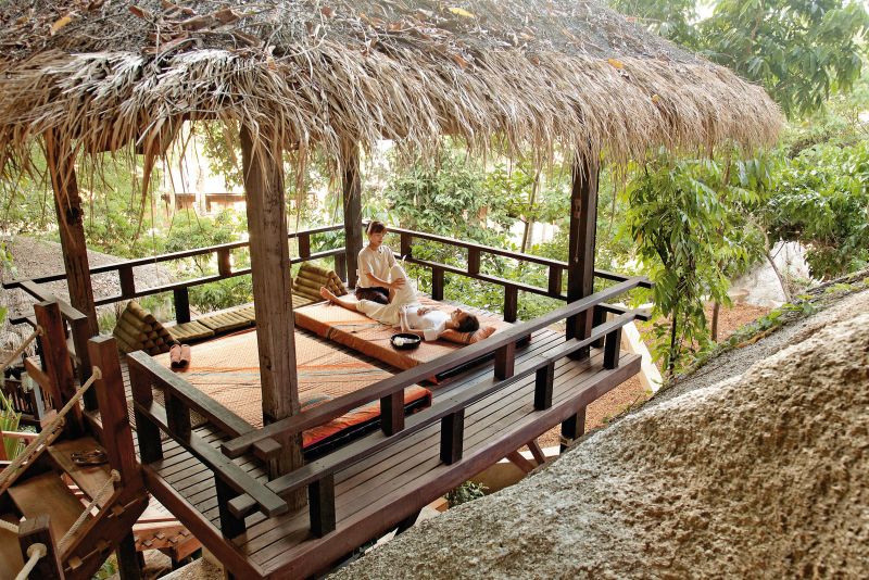 Spa Behandlung im Freien Eine Person, die eine Massage auf einer hölzernen Plattform unter einem Strohdach erhält, umgeben von üppiger Vegetation.