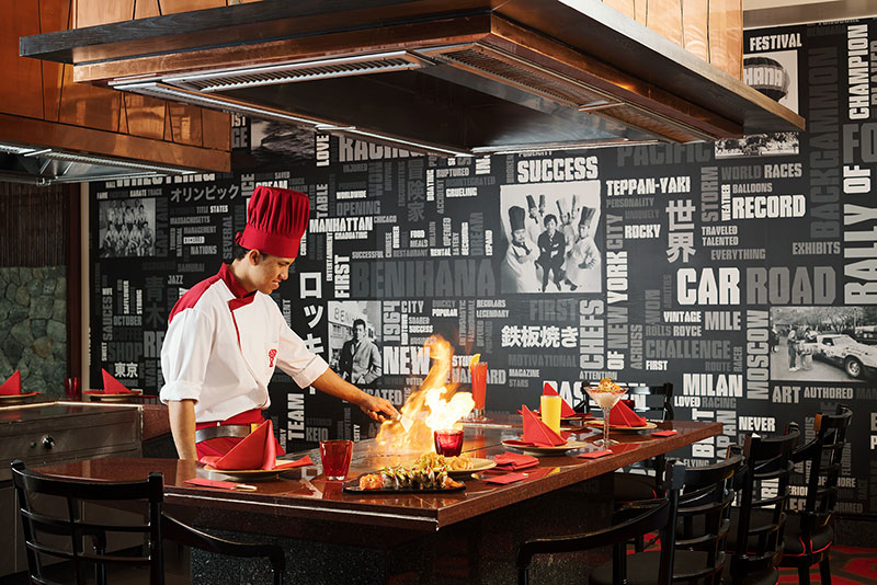 Benihana Restaurant Ein Koch in einem weißen Hemd mit rotem Kragen, roter Kochmütze und Schürze steht an einem Teppanyaki-Grill in einem Restaurant mit stilvollen, thematisch bedruckten Wänden. Er bereitet gerade eine Flambierung zu, wobei eine hohe Flamme über dem Grill emporzüngelt. Vor ihm auf dem Grill liegen verschiedene Speisen. Im Vordergrund ist ein ordentlich gedeckter Tisch mit roten Servietten und schwarzen Stühlen zu sehen.