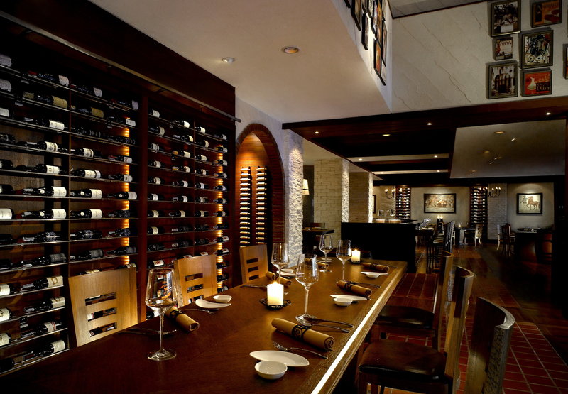 Italienisches Restaurant Brio Ein elegantes Restaurant mit gedimmter Beleuchtung, ausgestattet mit einem langen hölzernen Esstisch, der mit Geschirr, Weingläsern und Kerzen für das Abendessen eingedeckt ist. An der Wand befinden sich Regale voller Weinflaschen, und das Ambiente wirkt einladend und behaglich.