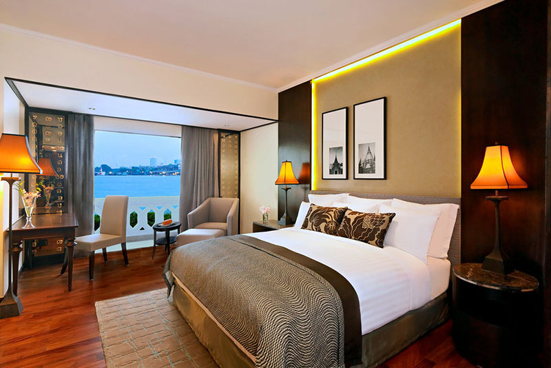 Deluxe Riverfront Zimmer Ein modern eingerichtetes Hotelzimmer mit einem großen Doppelbett, eleganten Nachttischlampen und einer Sitzgelegenheit am Fenster, das einen Blick auf die Wasserfront bietet.