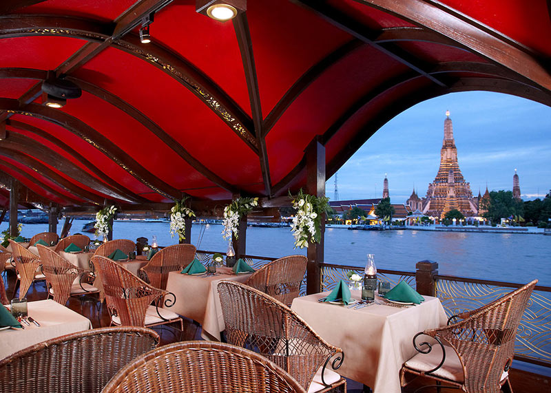 Manohra Dining Cruise Ein Blick von einer Restaurantterrasse mit rotem Sonnenschutz über den Fluss auf den beleuchteten Wat Arun Tempel in Bangkok während der Dämmerung. Vor den Tischen mit beige-braunen Tischdecken und geflochtenen Stühlen, einige mit grünen Servietten, erstreckt sich die ruhige Wasserfläche des Flusses.