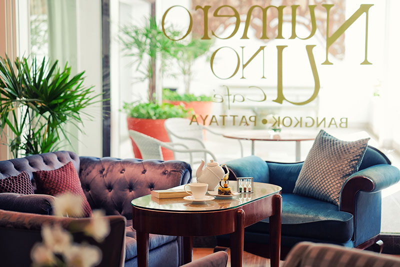 Numero Uno Cafe Gemütliches Café-Interieur mit einem dunkelblauen Samtsofa und einem Chesterfield-Sofa, einem Holztisch mit einer Teekanne und Tassen, dekorativem Licht und Pflanzen. Große Fenster mit Blick auf eine Terrasse befinden sich im Hintergrund, wobei der Schriftzug "LUNA PARK" spiegelverkehrt auf dem Glas steht.