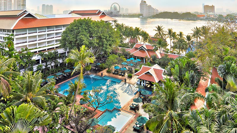 Resortüberblick Blick auf ein luxuriöses Resort mit roten Dächern, umgeben von tropischer Vegetation, mit mehreren Swimmingpools und einer attraktiven Gartenanlage, im Hintergrund ein Fluss und moderne Hochhäuser unter einem bedeckten Himmel.