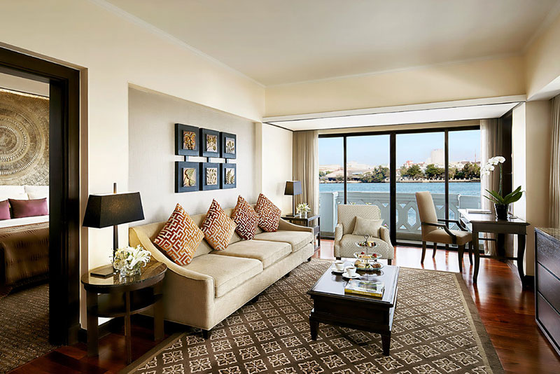 Wohnbereich der Riverfront Suite Ein stilvoll eingerichtetes Wohnzimmer mit einem großen, bequemen Sofa, dekoriert mit bunten Kissen. Im Hintergrund eröffnen sich durch bodentiefe Fenster Aussicht auf eine Wasserlandschaft. Ein eleganter Arbeitsbereich mit Schreibtisch und Stuhl befindet sich im rechten Bildabschnitt. Kunstwerke und dekorative Elemente vervollständigen das gehobene Interieur.