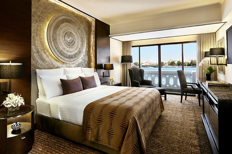 Riverfront Suite Ein luxuriöses Hotelzimmer mit einem großen, komfortablen Bett, auf dessen Kopfteil ein auffälliges, kreisförmiges Kunstwerk prangt. Elegante Beistelltische mit Lampen flankieren das Bett. Vor den bodentiefen Fenstern, die einen Blick auf eine Wasserfläche bieten, stehen zwei stilvolle Sessel und ein kleiner Tisch, perfekt für eine entspannte Aussicht. Die warme Farbgestaltung und die anspruchsvolle Einrichtung vermitteln ein einladendes Ambiente.