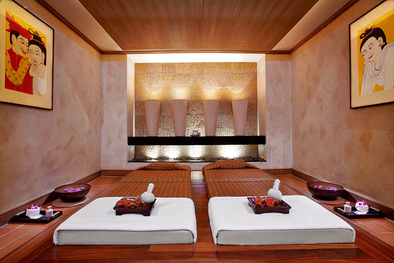 Anantara Spa Ein entspannend wirkender Massageraum mit drei weißen Massageliegen, dekoriert mit braunen Handtüchern und weißen Kissen. Vor den Liegen befinden sich hölzerne Schalen mit Blumen und Handtüchern. An der Wand hängen zwei großformatige Gemälde mit lachenden Figuren, und im Hintergrund ist eine Wand mit beleuchteten Nischen zu sehen. Der Raum hat ein warmes Ambiente mit Holzböden und einer Holzdecke.