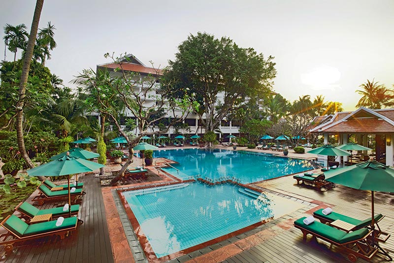 Swimmingpool Blick auf einen luxuriösen Poolbereich eines Resorts beim Sonnenuntergang, umgeben von tropischer Vegetation und Liegestühlen unter grünen Sonnenschirmen. Im Hintergrund ist ein mehrstöckiges Gebäude zu sehen.