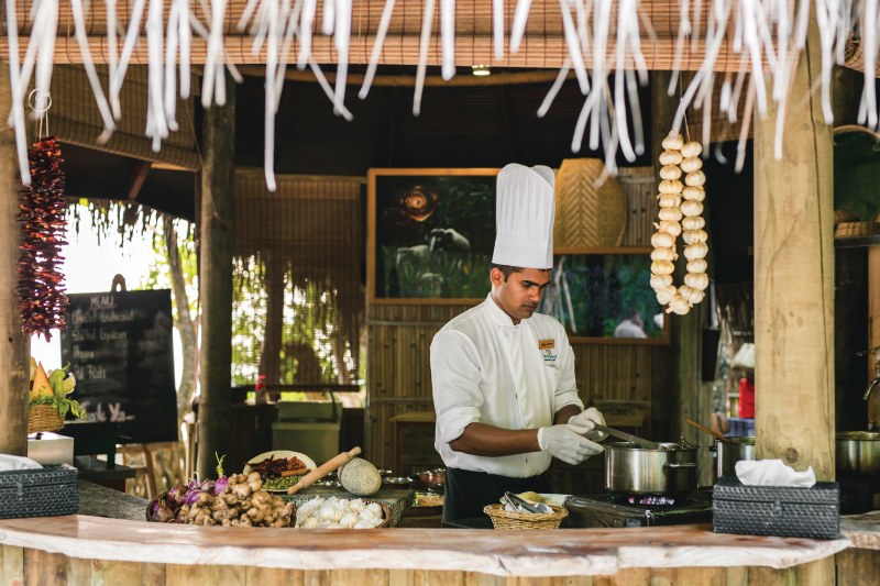 Ceylon Bliss Ein Koch in voller Berufsausstattung bereitet in einer rustikalen Außenküche Speisen zu. Er fokussiert sich darauf, in einem Topf zu rühren, der auf einem Tisch steht. Um ihn herum sind frische Zutaten wie Gemüse und Gewürze arrangiert. Aufhängende Zierobjekte und eine Tafel mit Menüvorschlägen tragen zum charmanten Ambiente bei.