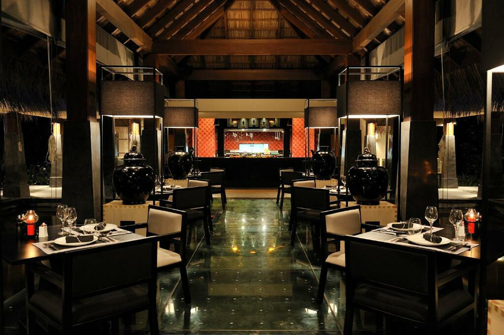 Gastronomie im Ayada Maldives Ein stilvolles und modernes Restaurant mit abendlichem Ambiente, großen schwarzen Zierurnen und elegant gedeckten Tischen, die auf einem glänzenden Boden ausgerichtet sind, im Hintergrund eine offene Küche.