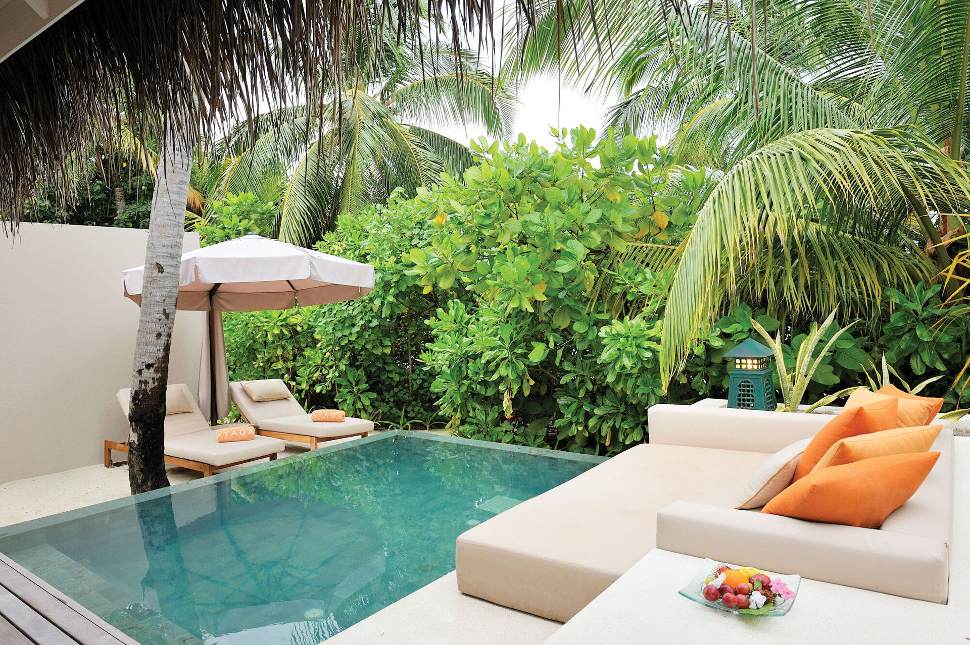 Privatpool im Ayada Maldives Ein luxuriöses Außenumfeld mit einem Pool, umgeben von üppigem Grün und Palmen. Zwei Sonnenliegen mit beigen Kissen und orangefarbenen Dekokissen stehen unter einem weißen Sonnenschirm neben dem Pool. Eine moderne, beige Lounge-Ecke mit passenden Kissen und einem Teller mit frischem Obst auf dem Tisch trägt zu einer entspannenden Atmosphäre bei.