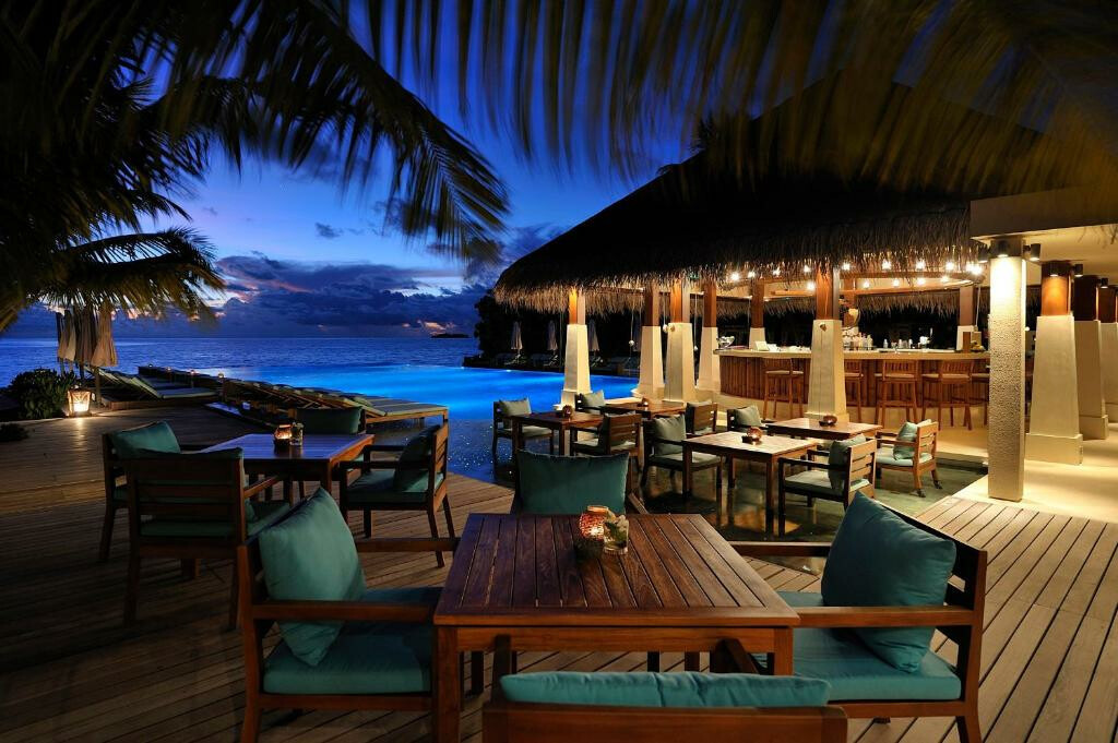 Abendstimmung im Ayada Maldives Alt-Text: Ein abendlicher Blick auf eine stilvolle Außenbar und ein Restaurant am Meer mit Holzmöbeln und türkisfarbenen Kissen. Beleuchtete Kerzen auf den Tischen schaffen eine gemütliche Atmosphäre. Im Hintergrund ist ein Schwimmbad zu sehen, das in blauem Licht erstrahlt, dahinter der Ozean und ein dramatischer Sonnenuntergangshimmel.