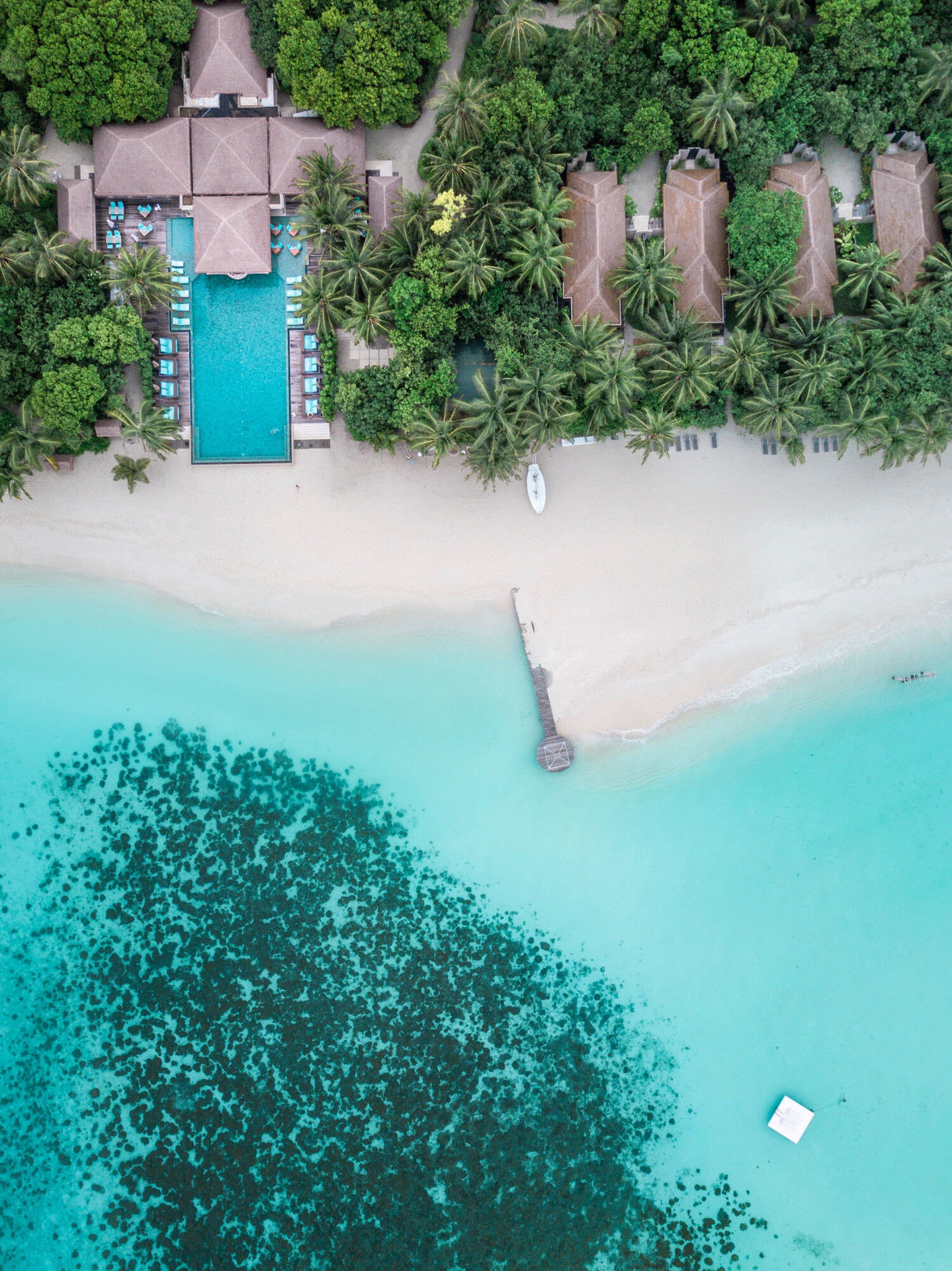 Das Ayada Maldives von oben Luftaufnahme eines luxuriösen Strandresorts mit einem großen zentralen Swimmingpool, umgeben von mehreren braun gedeckten Gebäuden, umgeben von üppiger grüner Vegetation. Ein weißer Sandstrand grenzt an kristallklares, türkisfarbenes Meerwasser, durchzogen von Korallenriffen. Ein Steg reicht ins Wasser hinein neben einem einzelnen Ruderboot am Ufer.