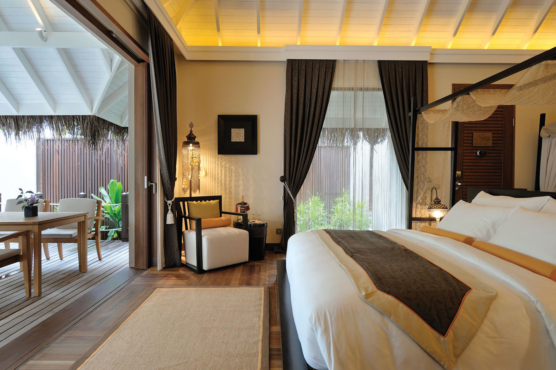 Wohnbeispiel des Ayada Maldives Ein elegantes Schlafzimmer mit einem großen Bett und weißen Bettwäsche im Vordergrund. Links sind eine Sitzgruppe und ein Esstisch auf einer Holzterrasse mit Blick auf grüne Pflanzen zu sehen. Das Zimmer ist reichhaltig beleuchtet und strahlt eine warme, einladende Atmosphäre aus.