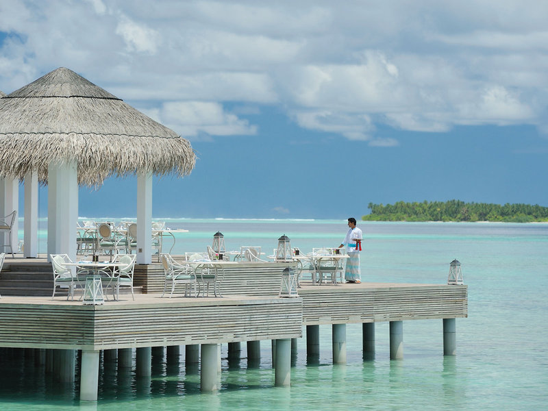 Restaurant Ein hölzerner Steg mit weißen Tischen und Stühlen führt zu einem Pavillon mit Strohdach vor einem türkisfarbenen Meer. Eine Person steht am Ende des Steges und blickt auf das Wasser. Im Hintergrund ist eine kleine Insel mit üppiger Vegetation zu erkennen.