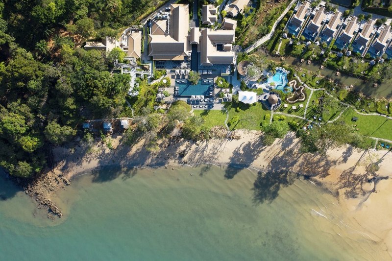 Blick auf das Banyan Tree Krabi Luftaufnahme eines luxuriösen Küstenresorts neben einem Sandstrand, umgeben von üppigen Bäumen, mit sichtbaren Gebäuden, Pools und angelegten Gartenanlagen.