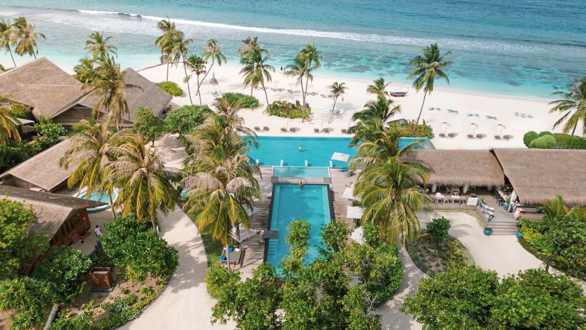 Ansicht von oben Ein Luftbild eines luxuriösen Strandresorts mit strohgedeckten Gebäuden, zwei Swimmingpools umgeben von üppigen Palmen und einer idyllischen weißen Sandstrandküste mit aufgestellten Liegestühlen am türkisblauen Meer.