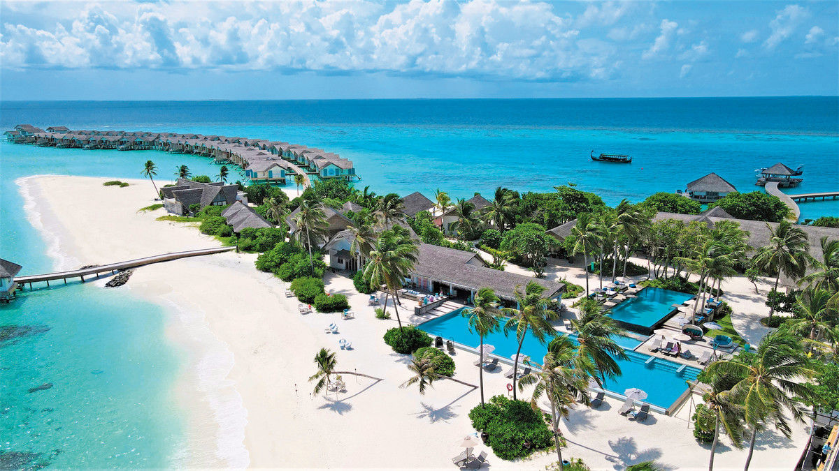 Außenansicht Ein luxuriöses Inselresort mit Überwasser-Bungalows und tropischen Villen, die sich an einem makellosen weißen Sandstrand entlangschlängeln. Mehrere Palmen säumen die Szenerie, während ein Swimmingpool als zentrales Element der Anlage fungiert. Im azurblauen Meer ist ein traditionelles Boot zu sehen, und die gesamte Atmosphäre vermittelt ein Gefühl maximaler Entspannung in einem exotischen Paradies.