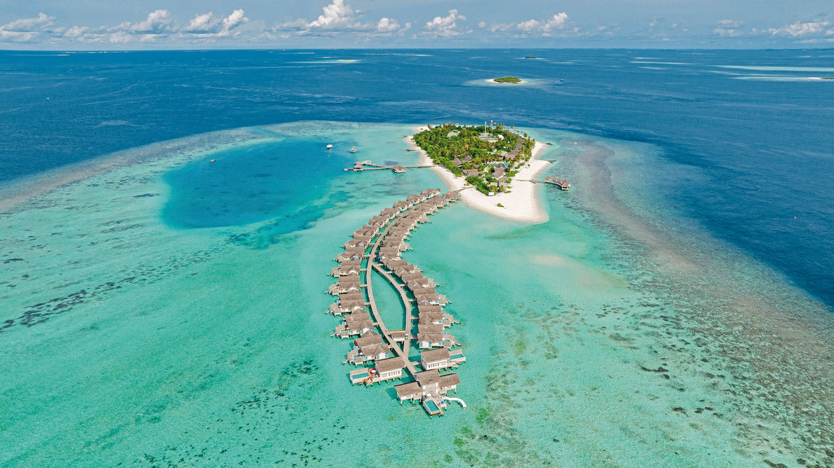 Draufsicht Luftaufnahme eines luxuriösen Inseldorfes auf den Malediven, mit einer Reihe von Überwasser-Bungalows, die sich in einem sanften Bogen über das türkisfarbene Meer erstrecken, umgeben von reichhaltigen Korallenriffen und weißem Sandstrand.
