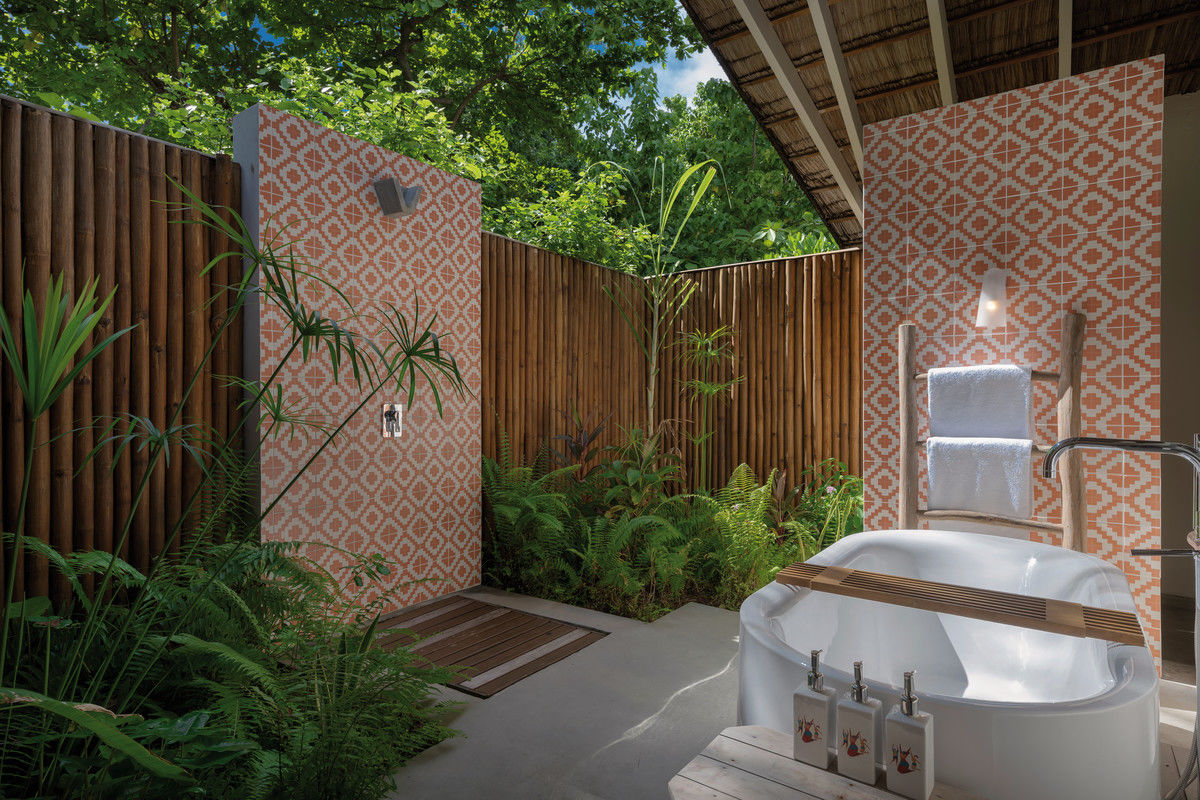 Outdoor Badezimmer Außenbereich einer Luxus-Spa-Suite mit einer freistehenden Badewanne, umgeben von tropischer Bepflanzung und Bambuszaun. Die Wände und der Boden sind in einem warmen, terrakottaähnlichen Muster gefliest. Handtücher hängen ordentlich an einer Wandhalterung neben der Wanne und natürliche Sonnenstrahlen durchdringen das blattreiche Umfeld, was eine entspannende Atmosphäre schafft.