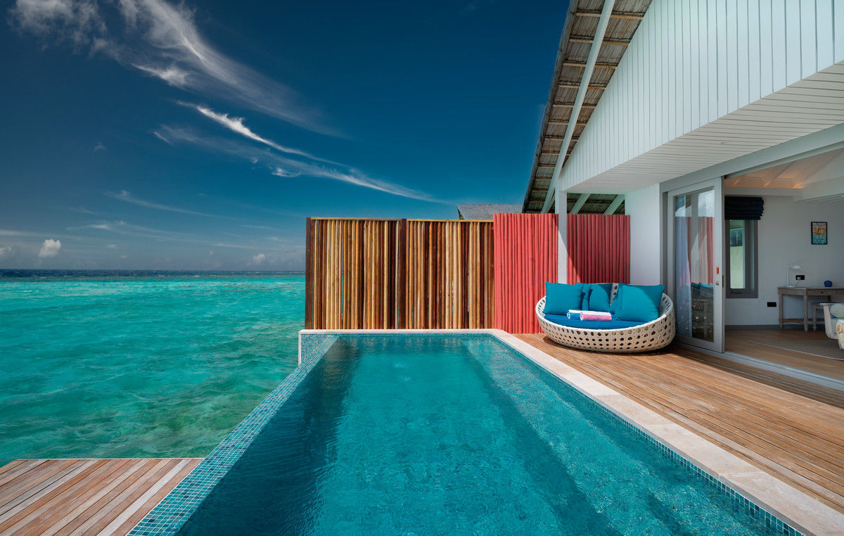 Privatpool Außenansicht einer luxuriösen Strandvilla mit privatem Pool, Holzterrasse und farbenfroher Lounge-Ecke, vor dem Hintergrund des türkisblauen Meeres unter einem klaren blauen Himmel mit wenigen Wolken.