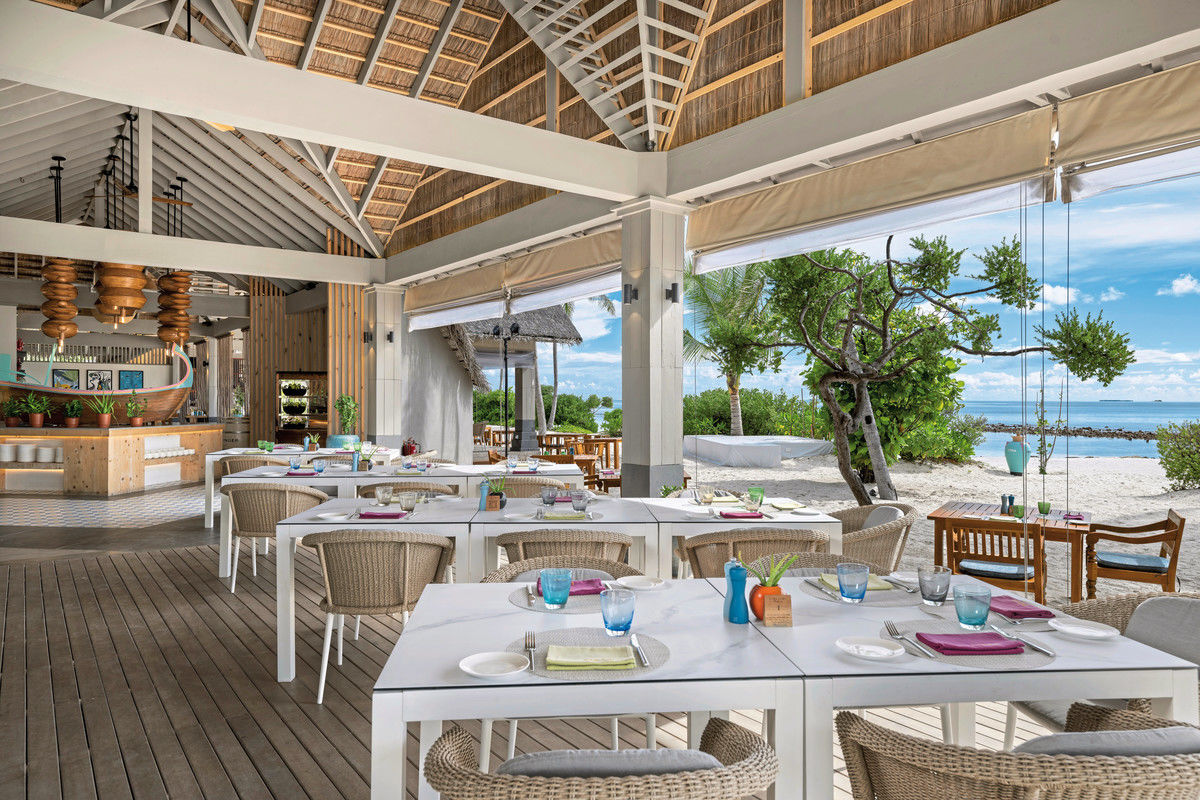 Restaurant Ein elegantes Strandrestaurant mit offenem Konzept, Holzmobiliar, weiß gedeckten Tischen und Dekoration in sanften Farbtönen. Im Hintergrund ist das Meer sichtbar und natürliche Vegetation umgibt die Terrasse.