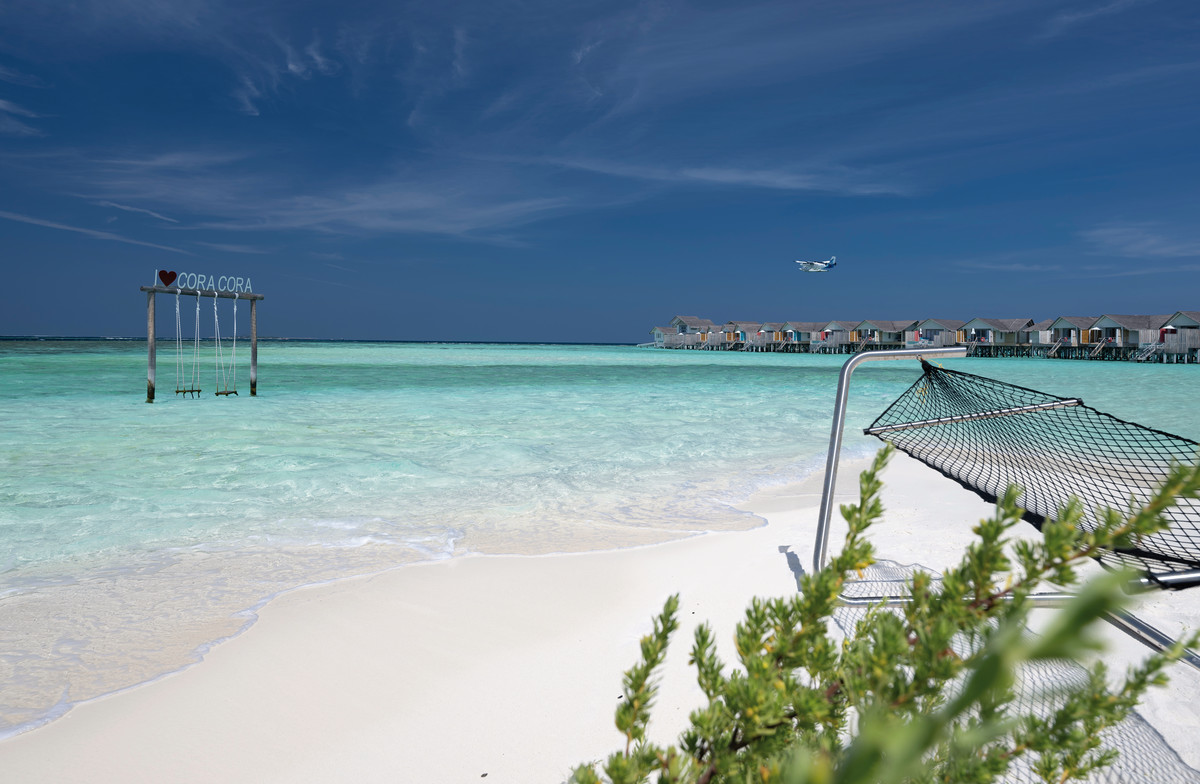 Strand Ein weißer Sandstrand mit kristallklarem, türkisfarbenem Wasser. Im Vordergrund ist eine Hängematte am Strand zu sehen. In der Mitte des Wassers steht ein hölzernes Gestell mit einer Tafel, auf der "CORA CORA" steht, und daran hängen Schaukeln. Im Hintergrund sind über dem Wasser auf Stelzen gebaute Bungalows zu erkennen und ein kleines Flugzeug ist im blauen Himmel sichtbar.