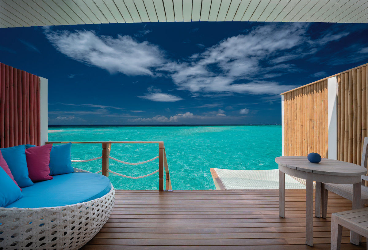 Terrasse Terrasse eines Luxus-Strandhauses mit einem runden Sofa mit blauen Kissen, einem hölzernen Tisch und Blick auf das kristallklare, türkisfarbene Meer unter einem weiten blauen Himmel mit wenigen Wolken.