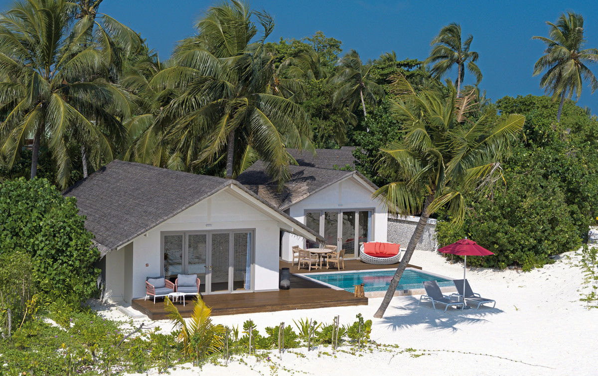 Wohnbeispiel von außen Ein luxuriöses Strandhaus umgeben von hohen Palmen auf weißem Sand. Vorne im Bild sehen Sie eine hölzerne Terrasse mit einem Pool, Sonnenliegen und einem roten Sonnenschirm, der Schatten spendet.