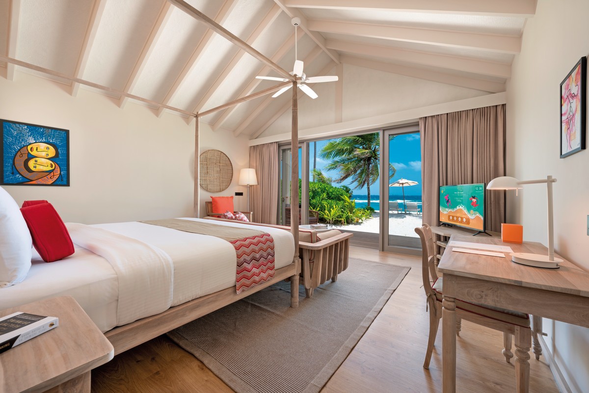 Wohnbeispiel Heller und stilvoller Hotelzimmer-Innenraum mit einem großen Bett, farbenfrohen Kunstwerken an den Wänden und einem direkten Blick auf den tropischen Strand durch eine Glasschiebetür.